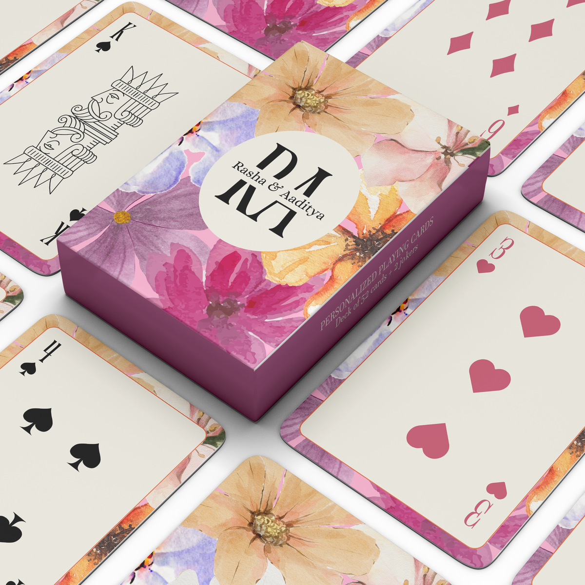 Personalised floral playing card set with pink and purple watercolor flowers, personalised initials and names on the box, and multiple playing cards arranged around it.